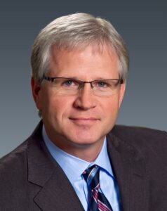 Eric C. Frimodt smiles in front of a gray background.